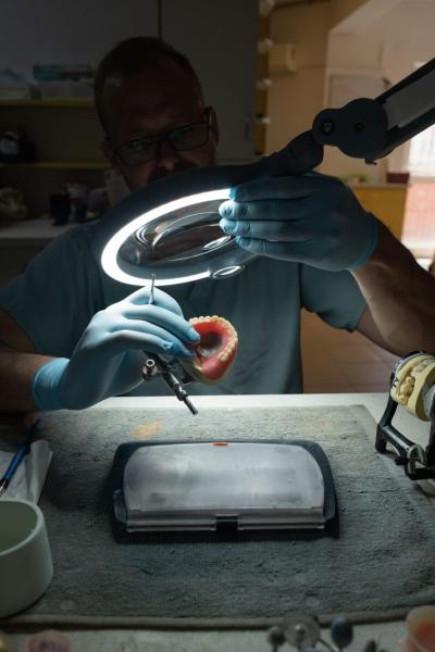 Ein Mensch mit Brille und Handschuhen arbeitet unter einer Leuchte. Er hält ein Modell eines Gebisses in der Hand, während er mit einem Werkzeug daran arbeitet. Der Hintergrund ist unscharf.
