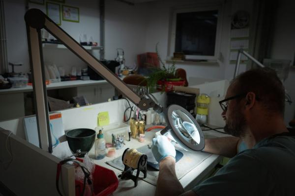 Ein Mann arbeitet konzentriert an einem Tisch unter einem großen, beleuchteten Vergrößerungsglas. Um ihn herum sind Werkzeuge und Materialien, die eine Werkstattatmosphäre erzeugen.