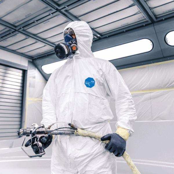 A person is wearing a white jumpsuit and a breathing mask. They are holding a hose with a spray device, ready to work in a bright, modern room.