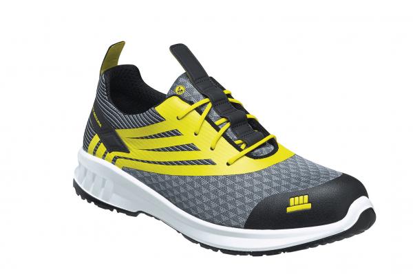 Der Schuh ist ein grauer Sneaker mit gelben Akzenten. Er hat ein sportliches Design, eine strukturierte Oberfläche und eine schwarze, verstärkte Zehenkappe. Die Schnürsenkel sind in der Mitte.