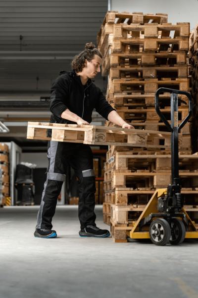 Eine Person in dunkelner Arbeitskleidung hebt eine Holzpalette an. Im Hintergrund sind viele gestapelte Holzpaletten zu sehen. Der Boden ist grau und die Umgebung wirkt industriell.