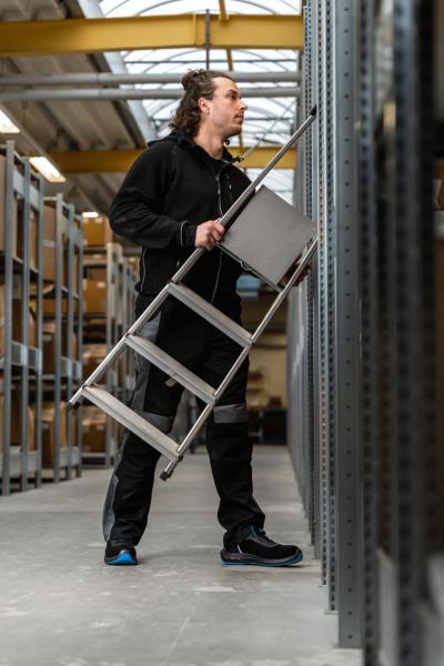 Ein Mann in schwarzer Arbeitskleidung steht in einem Lager und hält eine Aluminiumleiter in der Hand. Er schaut nach oben zu Regalen, um etwas zu erreichen. Die Umgebung wirkt industriell und gut organisiert.