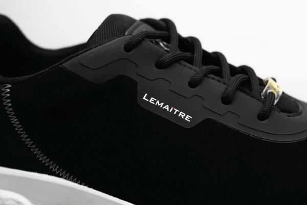 Das Bild zeigt einen schwarzen Sportschuh mit einem glatten Obermaterial. Die Schnürsenkel und eine weiße Gummisohle sind gut sichtbar. Ein kleines, silbernes Logo ist seitlich angebracht.