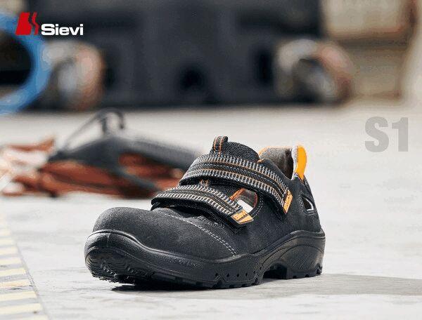 The image shows a black safety shoe with two Velcro straps. It is standing on a gray floor, with blurred work tools visible in the background.