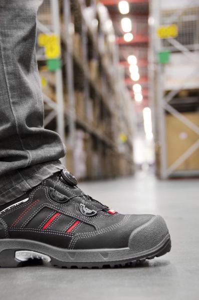Das Bild zeigt einen Fuß, der einen schwarzen Sicherheits-Schuh trägt, mit roten Akzenten. Im Hintergrund sind Regale mit Kisten und eine breite Lagerhalle zu sehen.