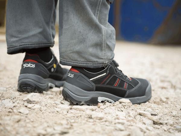 Das Bild zeigt die Füße einer Person in robusten, schwarzen Arbeitsschuhen auf einem steinigen Untergrund. Die Hosenbeine sind weit und grau, und es gibt farbige Akzente an den Schuhen.