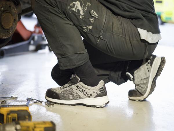 Eine Person sitzt auf dem Boden, trägt schwarze Arbeitshosen und graue, sportliche Schuhe. Werkzeuge liegen verstreut auf dem Boden, die Umgebung wirkt wie eine Werkstatt.