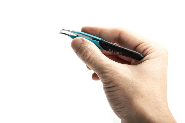 A hand is holding a pair of tweezers. The tweezers have a black handle with blue accents and a curved metal tip. The background is white and neutral.