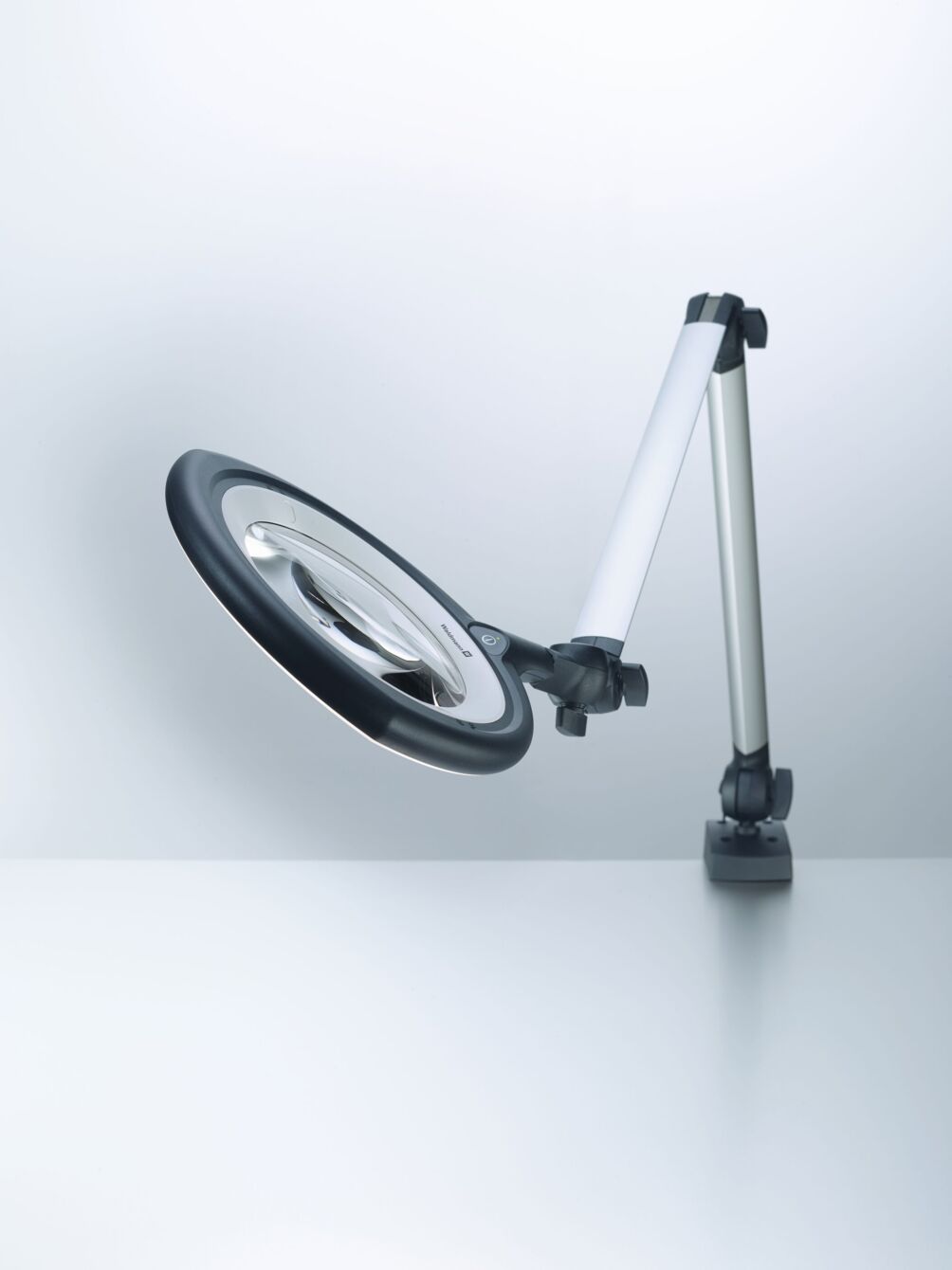 The image shows a flexible magnifying lamp with a large, round light ring. It stands on a stable base and has an adjustable arm that positions the lamp in various orientations.
