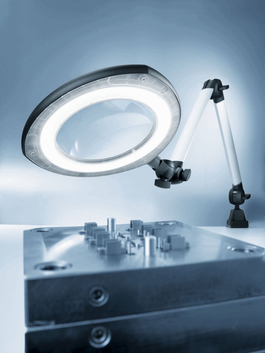 The image shows a magnifying lamp with ring lighting mounted on a table. Below it is a metallic component with various small tools or structures.