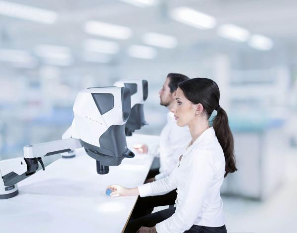 Two people in white shirts are sitting at a table in a modern room. In front of them are two large, futuristic-looking devices with screens. The room appears bright and tidy.