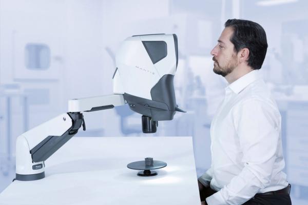 A man is sitting at a table, intently looking at a medical device with a movable arm. The device has a white surface and is located in a bright room.