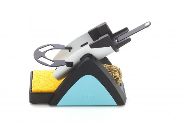 The image displays a modern soldering stand. It has a black base with a blue area, on which there is a yellow sponge. At the top, there is a holder for the soldering iron and a metal cleaning brush.