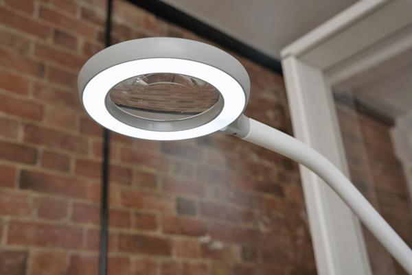 The image shows a white reading lamp with a circular LED ring and a magnifying lens in the center, mounted on a flexible arm. In the background, there is a brick wall.