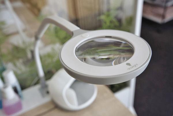 The image displays a bright magnifying glass with integrated light, mounted on a flexible arm. The background is blurred, featuring plants and another object that is not clearly identifiable.