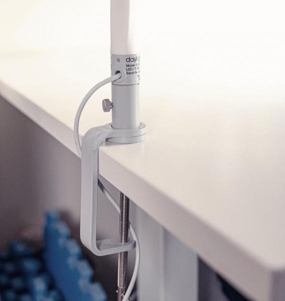 The image displays a white desk lamp attached to the edge of a table. It features a cylindrical shaft and a clamp for stabilization. The background appears bright and neutral.