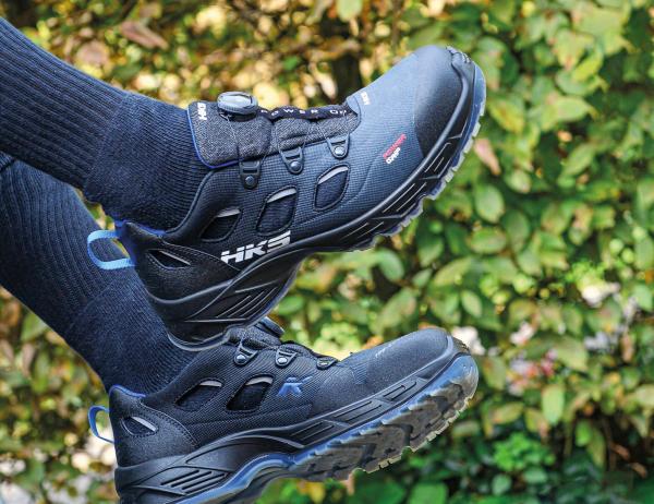 The picture shows a pair of black hiking boots with a sporty design. A person is wearing black socks, and in the background, there are green, blurred plants.