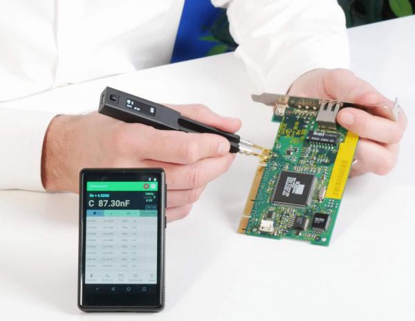 In the image, one hand is holding a circuit board while the other hand is using a measuring device. Next to the hand, there is a smartphone displaying values. The background is bright and neutral.