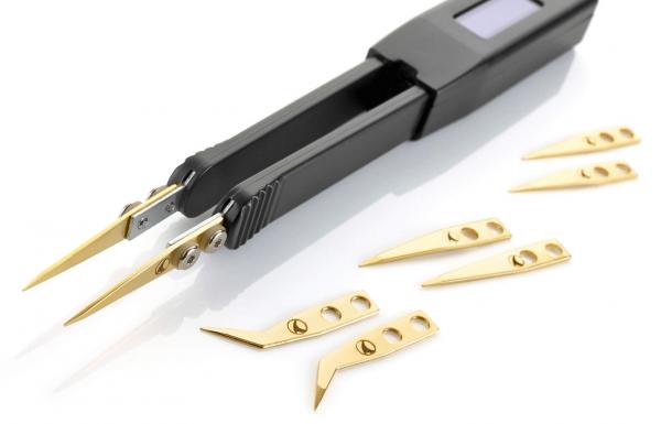 Auf dem Bild sind eine schwarze elektronische Pinzette mit zwei spitzen Greifarmen und mehrere kleine, goldene Schneideblätter zu sehen. Die Blätter sind unterschiedlich geformt.