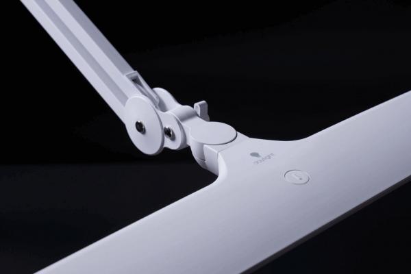 The image shows a modern, white desk lamp arm that is adjustable. The lamp has a simple, rectangular base and a light switch in the middle. The background is black.