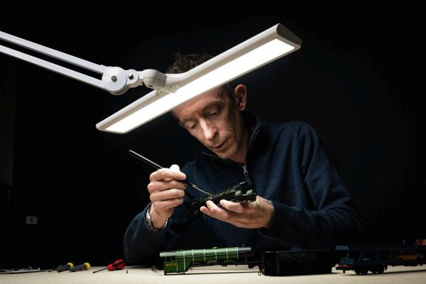A man is working intently on a small object under a bright lamp. He is holding a screwdriver and has a fine technique at hand. The background is dark.