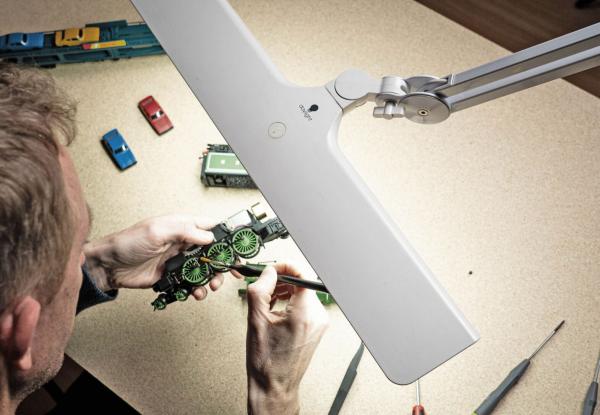 A person is working intently on a small model. In front of them is a bright lamp. Around them, small toy cars and tools are scattered.