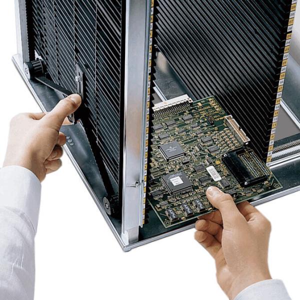 A person is holding a computer circuit board in their hand and is inserting it into a metallic case. In the background, cooling fins are visible. The focus is on the circuit board.