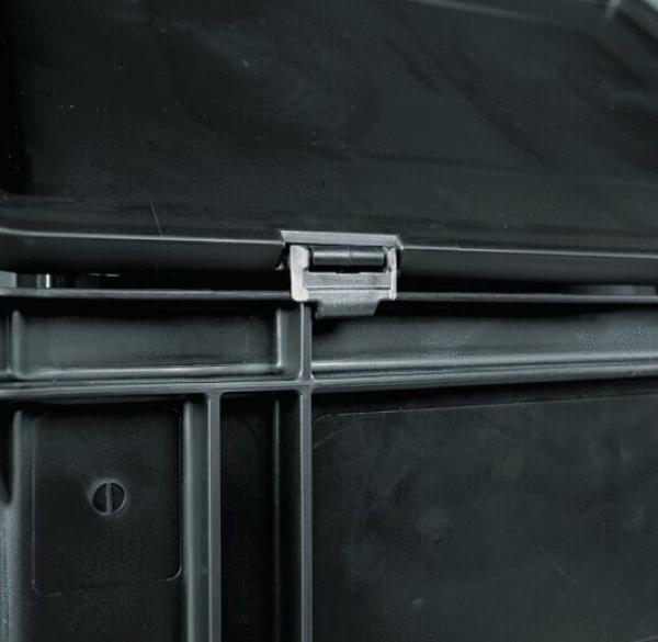 The image shows a dark, square plastic container. On the top, there is a clip closure that secures the lid. The bottom has some structural lines and indentations.