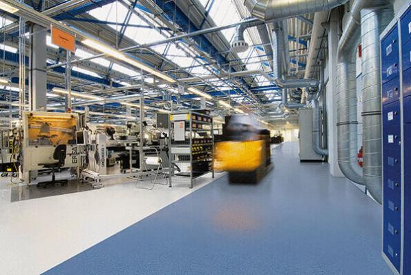 Das Bild zeigt eine moderne Fabrikhalle mit hoch Decken, blauen Stahlträgern und einem hellen Boden. Auf der linken Seite sind Maschinen und Regale, während ein Gabelstapler unterwegs ist.