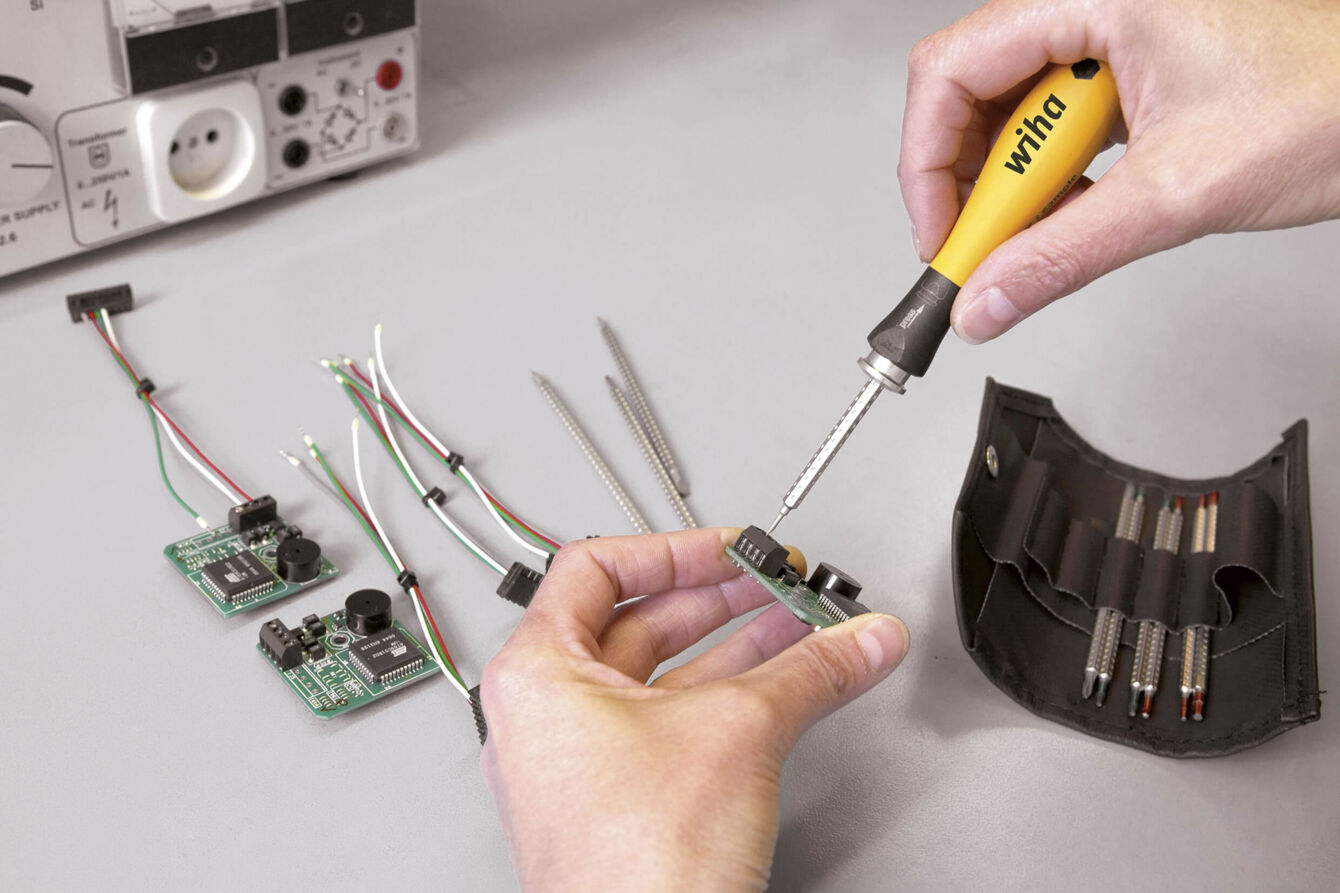 A hand holds a screwdriver over an electronic circuit board. Next to it, several cables and more circuit boards are lying around. In the background, a measuring device and a tool holder can be seen.