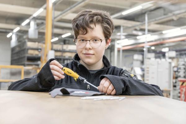 Ein Junge mit lockigen, braunen Haaren und einer schwarzen Jacke sitzt an einem Tisch. Er hält einen Schraubendreher und schaut konzentriert. Im Hintergrund sind Regale und Werkstattutensilien zu erkennen.