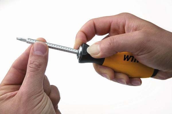 In the picture, there is a hand holding a screwdriver with a yellow handle. The tip of the screwdriver is pointing upwards, while a second hand carefully holds it steady.