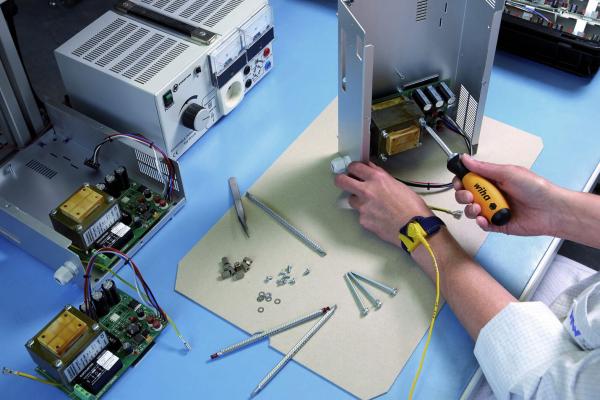 In the picture, you can see a work desk with technical equipment. A person is holding a screwdriver and working on an open case to repair electronic components. Around them, screws and tools are scattered.