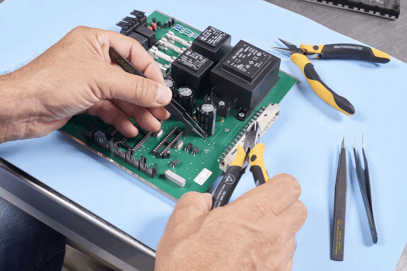 In the picture, there are two hands working on a green circuit board. The circuit board has various electronic components. Around the circuit board, there are tools such as pliers and tweezers.