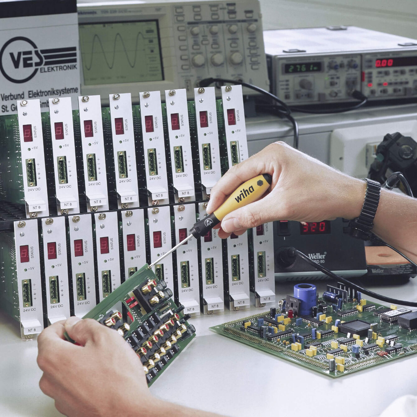 A hand holds a screwdriver and works on a circuit board. In the background are several devices with red displays, representing test and measurement instruments.