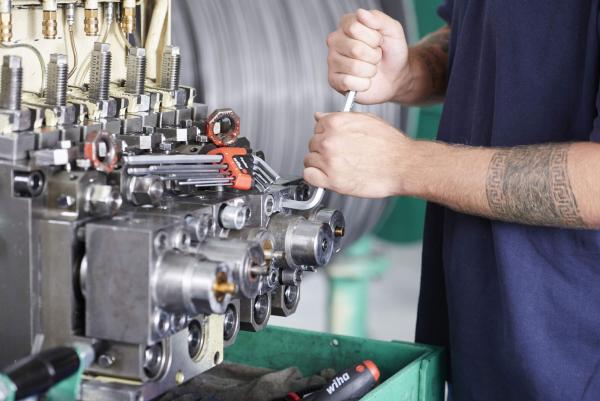 Ein Arbeiter in einem blauen Shirt repariert eine Maschinenkomponente. Er hält einen Schraubenschlüssel und arbeitet an einem Metallblock mit vielen Rohren und Verbindungen.