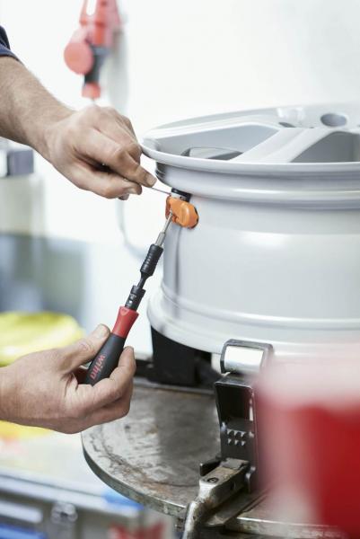 Ein händischer Werkstattvorgang: Eine Person verwendet ein Werkzeug, um an einer silbernen Felge zu arbeiten. Im Hintergrund sind Werkstattutensilien und eine helle Umgebung zu erkennen.