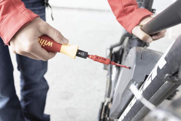 Eine Hand hält einen Schraubendreher mit rotem und gelbem Griff. Der Schraubendreher ist auf eine Schraube eines schwarzen Fahrrads gerichtet. Der Hintergrund ist hell und unscharf.