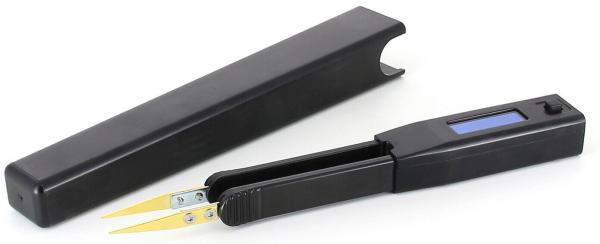 Das Bild zeigt ein schwarzes Gerät mit zwei goldenen Zangen, das geöffnet ist, und einem rechteckigen Gehäuse. Neben dem Gerät liegt eine schwarze Kappe oder Schutzhülle.