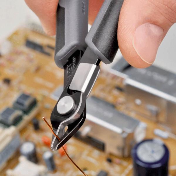 A hand holds a wire cutter pliers gripping a thin copper wire. In the background, a circuit board with various components is visible.