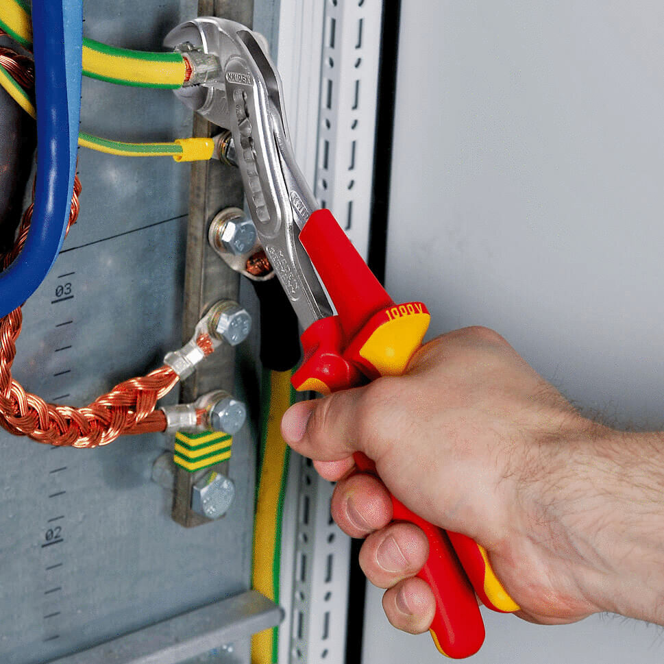 In the picture, you see a hand holding an adjustable wrench. The wrench has red and yellow handles and is used to secure cables on a metal casing.