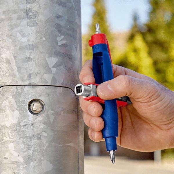 Eine Hand hält ein farbenfrohes Werkzeug, das aussieht wie ein Stift mit einer kleinen Ratsche. Es wird an einem runden, metallischen Pfosten verwendet, um eine Schraube zu befestigen.