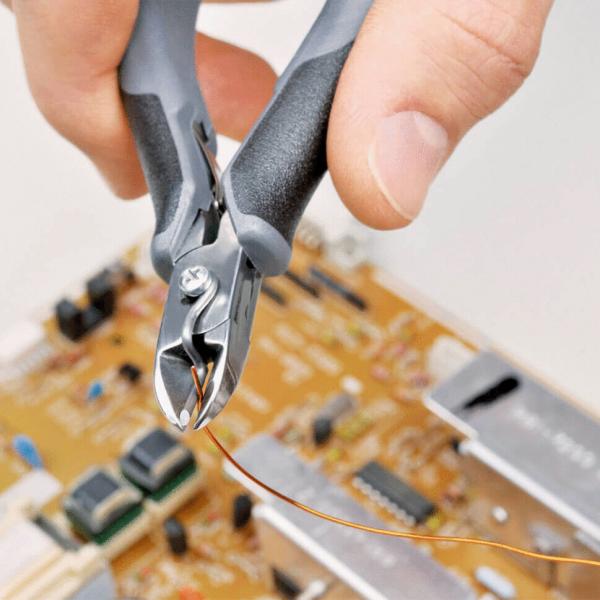Eine Hand hält eine Zange, die dünnen Kupferdraht schneidet. Im Hintergrund ist eine Leiterplatte mit verschiedenen elektronischen Bauteilen und einer goldenen Oberfläche zu sehen.
