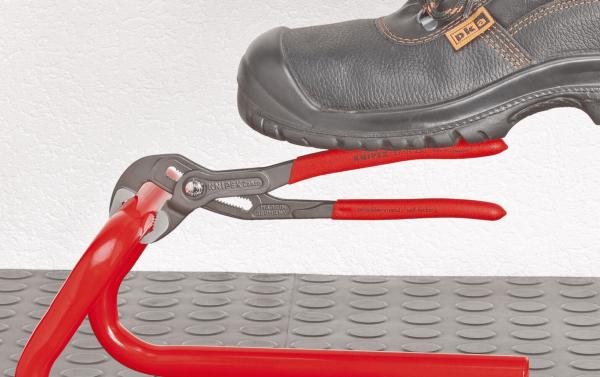 A black work shoe is standing on a red pliers, which is lying on a gray rubber floor. The pliers are open and have red handles. The wall in the background is white.
