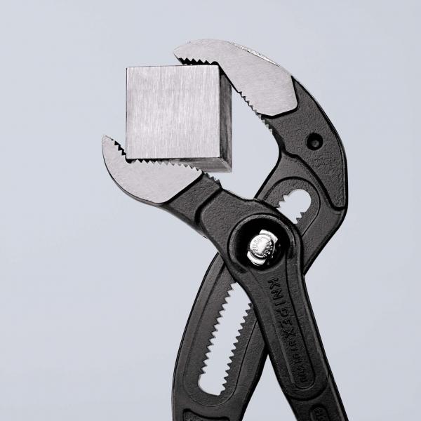 Das Bild zeigt eine schwarze Zange, die einen kleinen, quadratischen Metallblock festhält. Der Hintergrund ist hell und neutral gehalten.
