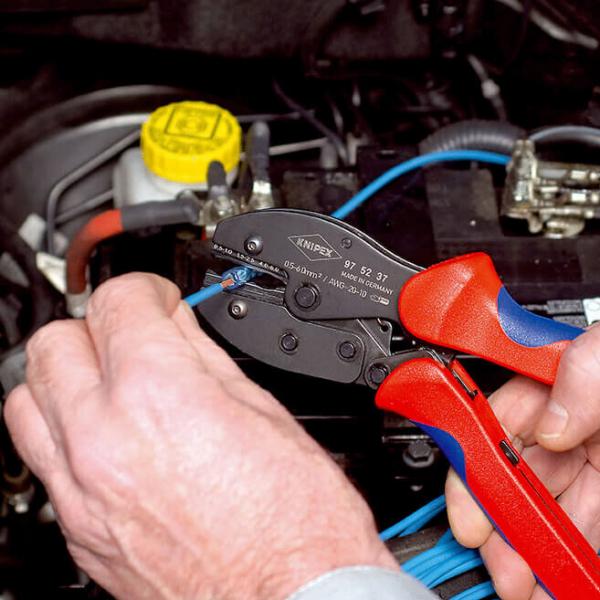 A hand holds a wire cutter with red and blue handles while working on a blue cable leading to a car component. In the background, a yellow container is visible.