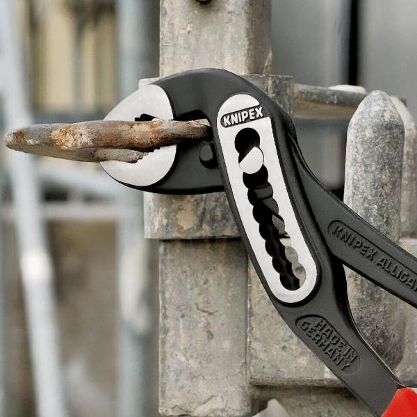 The image shows a Knipex pliers attached to a metal fence. The pliers are gripping a rusty key that is slightly protruding from the fence.