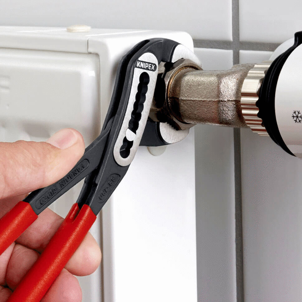 A hand is holding pliers on a heating pipe. The pipe is made of metal and mounted on a white radiator. The pliers have black and red handles.