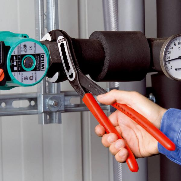 In the picture, there is a hand holding a water pump pliers with red handles. It is being attached to a large, black pipe with a green pump and a pressure gauge.