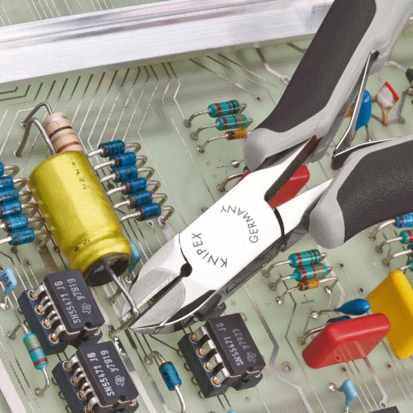 Das Bild zeigt ein Leiterplattenlayout mit verschiedenen elektrischen Komponenten. In der Mitte liegen flache, schwarze Bauteile und ein gelber Kondensator. Eine Zange greift eine der Komponenten.